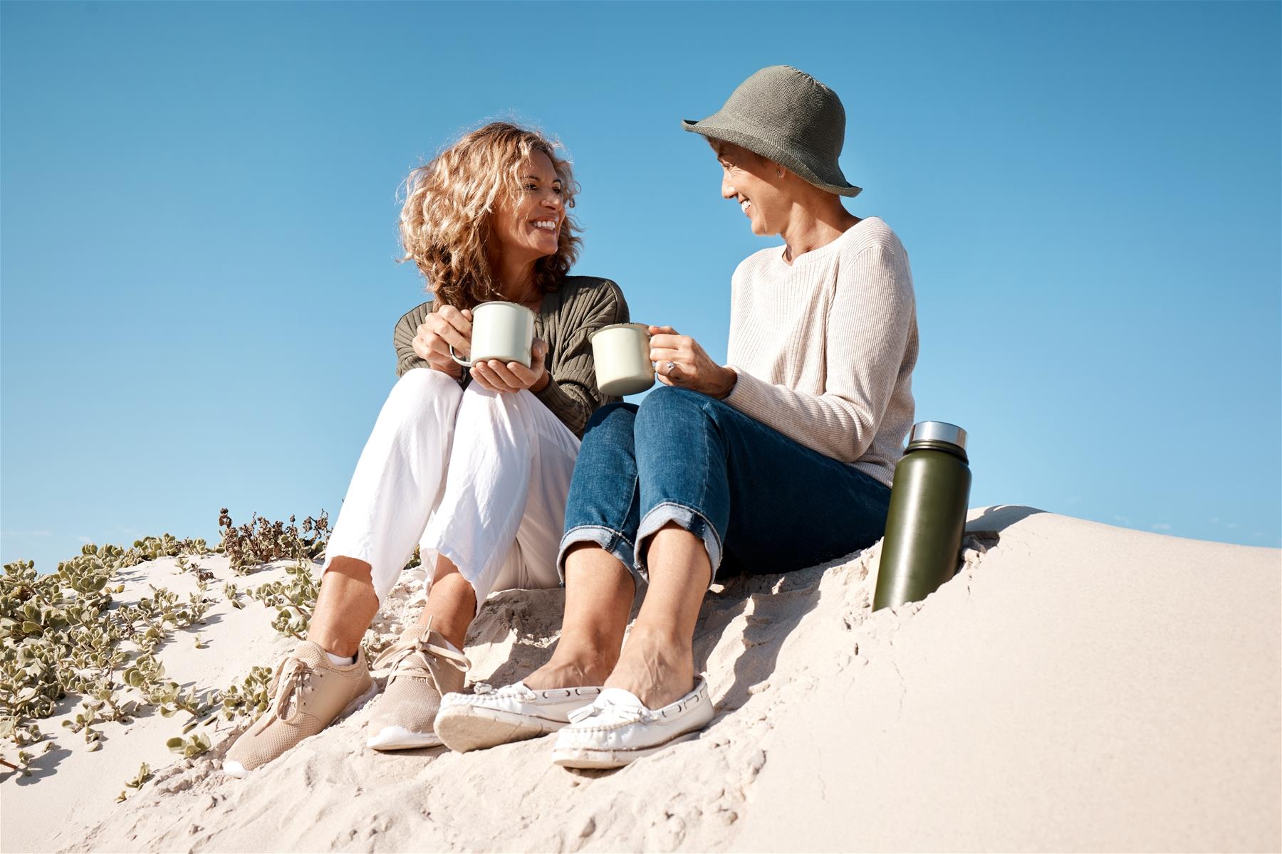 Two ladies sit on a sand dune and share a cuppa.