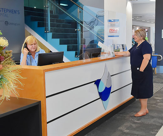 A woman answers the phone at the St Stephen’s Hospital reception desk, while a staff member waits.